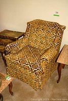 Front angled view of vintage fabric upholstered arm chair with distinct brown and gold pattern, showing wear and rip on fringe.