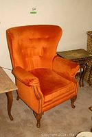 Front left-side view of the orange velour upholstered arm chair showing tufted backrest, padded arms, wooden feet and nail head trim along the armrest edges.