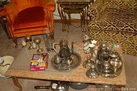 Wide view of assorted silverplate pieces including teapots, trays, candlesticks, brass oil lamps, sugar and cream pots on trays placed on table and floor with surrounding furniture.