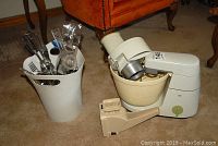 Photo of Braun stand mixer with mixing bowl and juicer attachment beside white container holding utensils on carpeted floor.