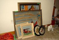 Photo showing the group of seven framed needlepoint art pieces stacked and leaning against a wall, with two oval frames, two small square frames with antique cars, and three larger rectangular framed pieces behind them.