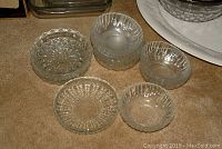Five clear glass bowls with decorative ribbing and floral patterns on beige carpet. Used for display of glassware patterns.