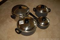 Four grey cooking pots with black handles and stainless steel lids on a carpeted floor.