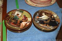 Two stacks of Norman Rockwell collector plates with illustrations of children and adults in vintage home scenes, placed on a blue cloth.