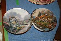 Photo of two collector plates showing detailed cottage scenes with flowers and swans, gold trim on edges.