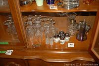 Photo of assorted crystal stemware glasses, two pairs of salt and pepper shakers, ceramic egg coddlers, and small glass vases arranged inside a wooden hutch