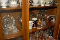 Display cabinet shelf with metal fish platter, Sandringham stamped plates, teacups, cake plate, oil lamps, and salt and pepper shakers