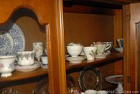 Teacups and saucers displayed inside a wooden cabinet, multiple patterns and designs visible