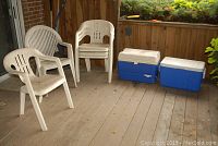 Eight white plastic lawn chairs with armrests and two blue and white Igloo coolers on wooden porch floor, near brick wall.