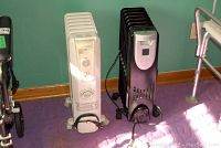 Two electric portable heaters, one white Noma and one black Bionaire, placed side by side on purple carpet near green wall.