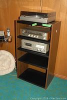 Photo of vintage stereo system components arranged in the faux wood cabinet on blue carpeted floor with wood panel wall background.