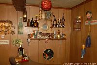 Wide view of bar corner with multiple shelves holding assorted small bottles, shot glasses, and metal wall art including a large bottle cap shaped sign and a round decorative plate