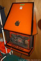 Front view of the vintage electric wall fireplace showing the orange and black finish with a metal ship decoration on the slanted top.