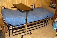 Full view of adjustable medical bed with metal frame, blue mattress and overbed tray in lowered position