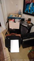 Photo showing multiple office items including a computer tower, paper shredder, briefcases, and a box of stationery.