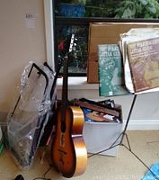 Acoustic guitar, black metal keyboard stand, musical books, and case visible by window with greenery outside. Guitar has sunburst finish.