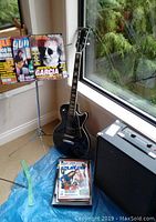 Black Harmony electric guitar leaning against window, practice amp behind, tripod music stand holding magazines