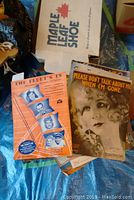 Vintage sheet music with period illustrated covers visible, some titles readable, set on a surface with Maple Leaf Shoe paper in background.