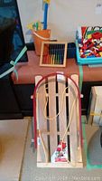 Wooden jab sled with red runners at front, orange bucket, toy abacus, box with markers, and shovel visible on brown surface