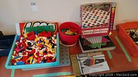 Top view of blue tray filled with assorted loose Lego pieces next to a Chinese Checkers box, red bucket, and other items on a table.