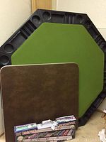 Octagonal poker table with green felt surface, black chip holders around the edge, folded rectangular card table, and boxed poker set in front.