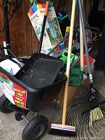 Photo showing black plastic wheelbarrow, rake, broom and other garden tools in garage setting.