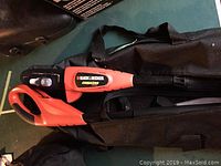 Black and Decker cordless pole saw resting on its black carrying bag, showing ergonomic handle and orange-black design.