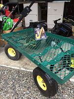 Green metal garden cart with mesh bed seen from an angle showing yellow wheels and a packaged grip trailer wheel lock sitting on the cart bed.