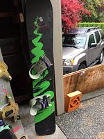 Full snowboard with bindings leaning against wall outside, black with green and red graphics, white and purple bindings attached.
