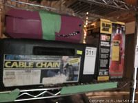 Photo showing Black Jack 12 ton hydraulic bottle jack box, two tire chain cases (one black labeled Cable Chain), partially covered purple case, on metal shelf.
