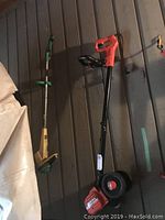 Photo showing two tools leaning against a wall: an orange and black wheeled edger and a green and yellow weed eater trimmer