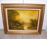 Frontal view of the framed oil painting showing the house on a river and the brown gold wooden frame.