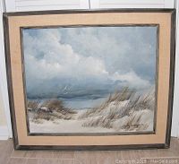 Full view of large framed beach seascape oil painting showing sand dunes with grass under a cloudy sky and sea in the distance.