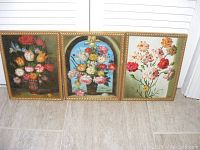 Three framed floral paintings placed side by side on floor, showing decorative flowers and gold frames