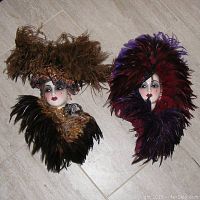 Both porcelain lady face masks side by side on floor, showing full feathered hats and collars.