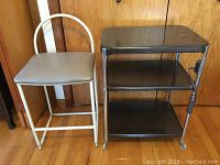 Padded island stool with white metal frame and gray vinyl seat sitting beside a dark metal table with three shelves, wheels, and an electric outlet on the side.