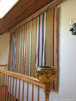 Woven Mexican blanket hung on wall showing colorful vertical stripes with tan background and wooden railing in front. Clay pot positioned on wooden post in foreground.