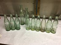 Nine vintage Coca-Cola glass bottles of varied designs arranged in a row on a white surface showing embossed logos and different bottle shapes.