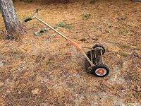 Full view of one old-fashioned push reel lawn mower with wooden handle and black grips, placed on dry grass.