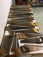 Photo shows a lineup of 11 old wooden hand saws placed on a white table, varying handle shapes and blade sizes.