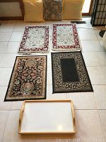 Overview of four small oriental rugs (two light colored, two dark) arranged on floor with four throw pillows propped against wall and wooden serving tray in foreground