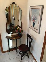 Corner view showing antique piano chair, half-moon entry table, large wall mirror with fluted edges, vase with silk flowers, and framed floral art print on adjacent wall.