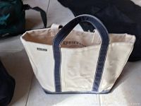 Beige canvas tote bag with dark blue handles and bottom visible on tiled floor, showing size and handle detail.