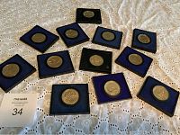 Twelve American Revolution Bicentennial medals displayed on a white lace cloth surface, each in individual display cases with blue or green felt lining.