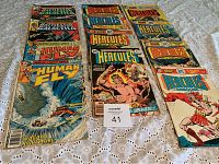 Comic books showing Battlestar Galactica, The Human Fly, Hercules Unbound issues arranged on cloth surface.