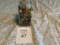 Front view of glass jar containing various marbles and coins on a white lace tablecloth with number 47 auction tag in front.