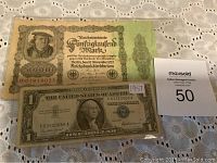 Photo showing both banknotes: the German 50000 Mark note on top and the US One Dollar silver certificate below, placed on a lace tablecloth.