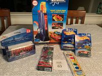 Lot of toys on table including Hot Wheels Fusion Factory box, Marvel Super Heroes Power Disc Capsule, Hot Wheels cars, gel pens, and wooden rattle