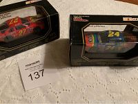 Photo of both NASCAR model cars number 24 and 27 in original packaging, boxed, placed on fabric surface with MaxSold lot number card 137.
