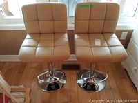 Pair of beige faux leather upholstered bar stools with square tufted pattern, chrome stands, and round flat bases, placed on a hardwood floor near a window and wall outlet.